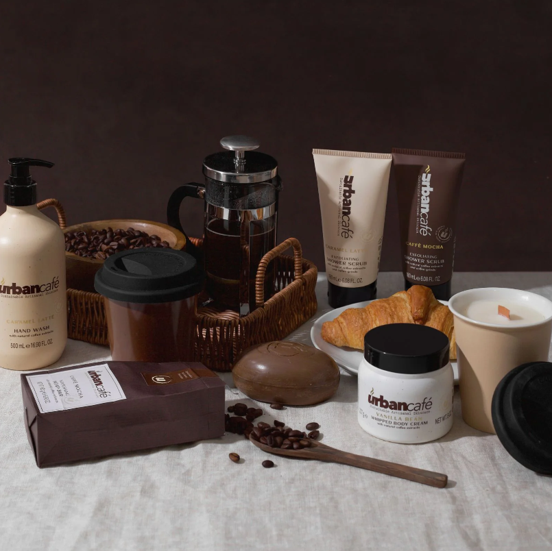 Die Handseife „Urban Caffé Vanilla“ (500 ml) von Somerset Toiletry ist auf einer braunen Fläche neben Kaffeebohnen, Croissants, einer French Press, einer Kerze und Kaffeebechern zum Mitnehmen angeordnet, alles vor einem dunkelbraunen Hintergrund.