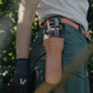 Eine Person in grüner Hose und Handschuhen steht im Freien, am Gürtel befestigt eine Niwaki | „Long Holster“ Lederschutzhülle für große Scheren (28,4 × 8,2 cm) von Niwaki, und hält eine Gartenschere in der Hand, während im Hintergrund Sonnenlicht durch die Blätter scheint.