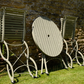 Das Harrod Horticultural Bistro Set 'Darsham' - ein klappbarer Gartentisch mit 2 Stühlen in Salbeigrün - bringt Charme in Ihren Garten, aufrecht im grünen Gras neben einer sonnenbeschienenen Steinmauer.