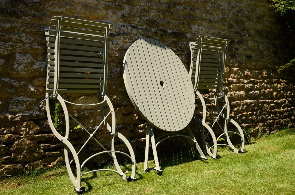 Das Harrod Horticultural Bistro Set 'Darsham' - ein klappbarer Gartentisch mit 2 Stühlen in Salbeigrün - bringt Charme in Ihren Garten, aufrecht im grünen Gras neben einer sonnenbeschienenen Steinmauer.