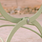 Nahaufnahme eines salbeigrünen Stuhl- oder Tischbeins aus Metall mit gebogenen Stützen und sichtbarer Schraube, Teil des Harrod Horticultural Bistro Set 'Darsham' Klappbarer Gartentisch mit 2 Stühlen in Salbeigrün, vor einem unscharfen Hintergrund im Freien.