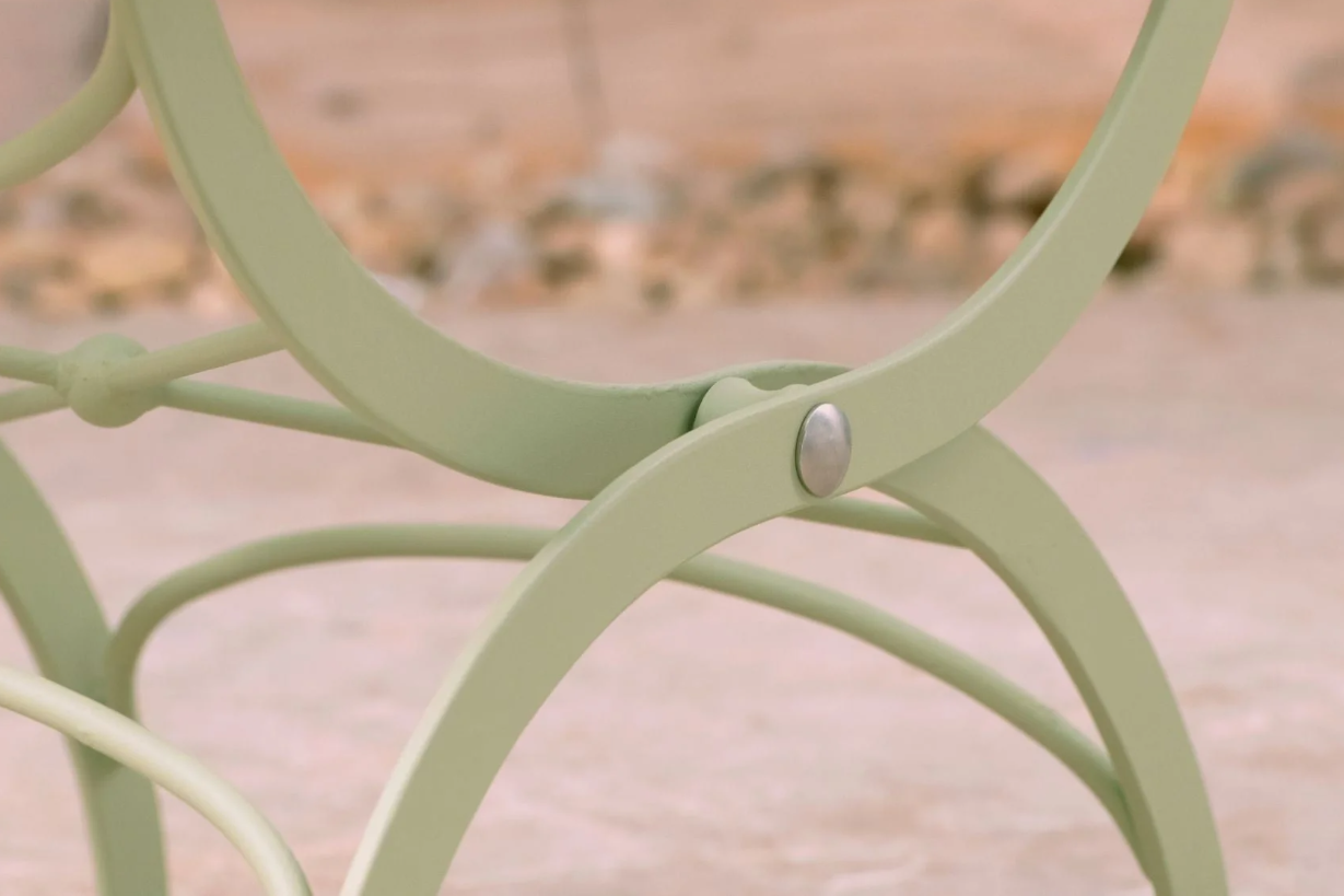 Nahaufnahme eines salbeigrünen Stuhl- oder Tischbeins aus Metall mit gebogenen Stützen und sichtbarer Schraube, Teil des Harrod Horticultural Bistro Set 'Darsham' Klappbarer Gartentisch mit 2 Stühlen in Salbeigrün, vor einem unscharfen Hintergrund im Freien.