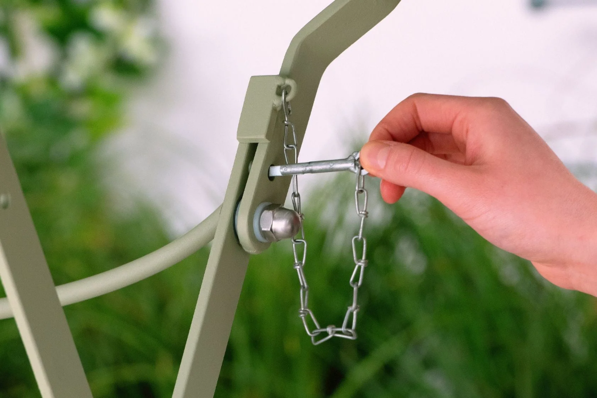 Eine Hand setzt einen Metallstift an einer Kette in den Rahmen des Harrod | Bistro Set 'Darsham' Klappbarer Gartentisch mit 2 Stühlen in Salbeigrün von Harrod Horticultural ein, wobei das Grün im Hintergrund sanft verschwimmt.
