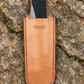 Ein Werkzeug mit schwarzem Griff ist teilweise in das hellbraune Niwaki | „Saw Holster“, einer hochwertigen Lederschutzhülle für die Klappsäge (26,5 × 8,4 cm), die mit dem Niwaki-Logo versehen ist und auf einer strukturierten Felsenoberfläche ruht – ein Must-have für die stilvolle und langlebige Aufbewahrung Ihrer Gartenwerkzeuge.
