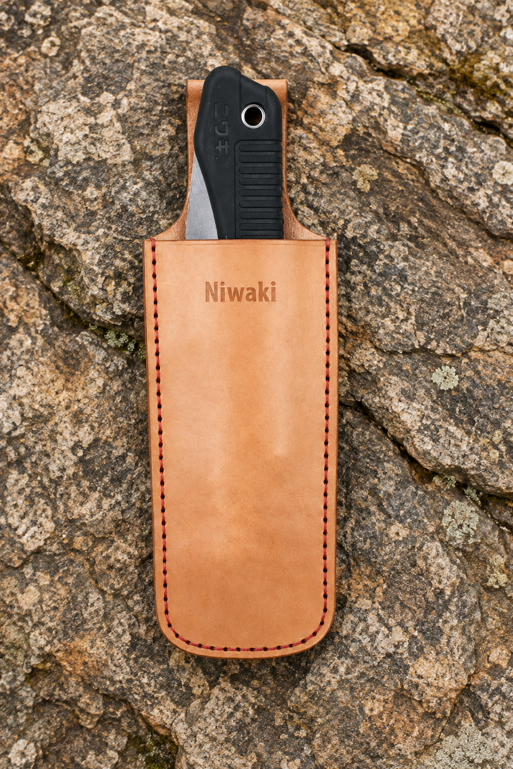 Ein Werkzeug mit schwarzem Griff ist teilweise in das hellbraune Niwaki | „Saw Holster“, einer hochwertigen Lederschutzhülle für die Klappsäge (26,5 × 8,4 cm), die mit dem Niwaki-Logo versehen ist und auf einer strukturierten Felsenoberfläche ruht – ein Must-have für die stilvolle und langlebige Aufbewahrung Ihrer Gartenwerkzeuge.