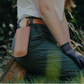 Eine Person kniet im Gras und trägt eine grüne Hose, braune Stiefel, schwarze Handschuhe sowie eine hochwertige Lederschutzhülle „Saw Holster“ von Niwaki für eine Klappsäge (26,5 × 8,4 cm) am Gürtel. Im sanft unscharfen Hintergrund sind hohes Gras und Bäume zu sehen.