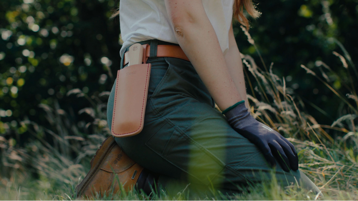Eine Person kniet im Gras und trägt eine grüne Hose, braune Stiefel, schwarze Handschuhe sowie eine hochwertige Lederschutzhülle „Saw Holster“ von Niwaki für eine Klappsäge (26,5 × 8,4 cm) am Gürtel. Im sanft unscharfen Hintergrund sind hohes Gras und Bäume zu sehen.