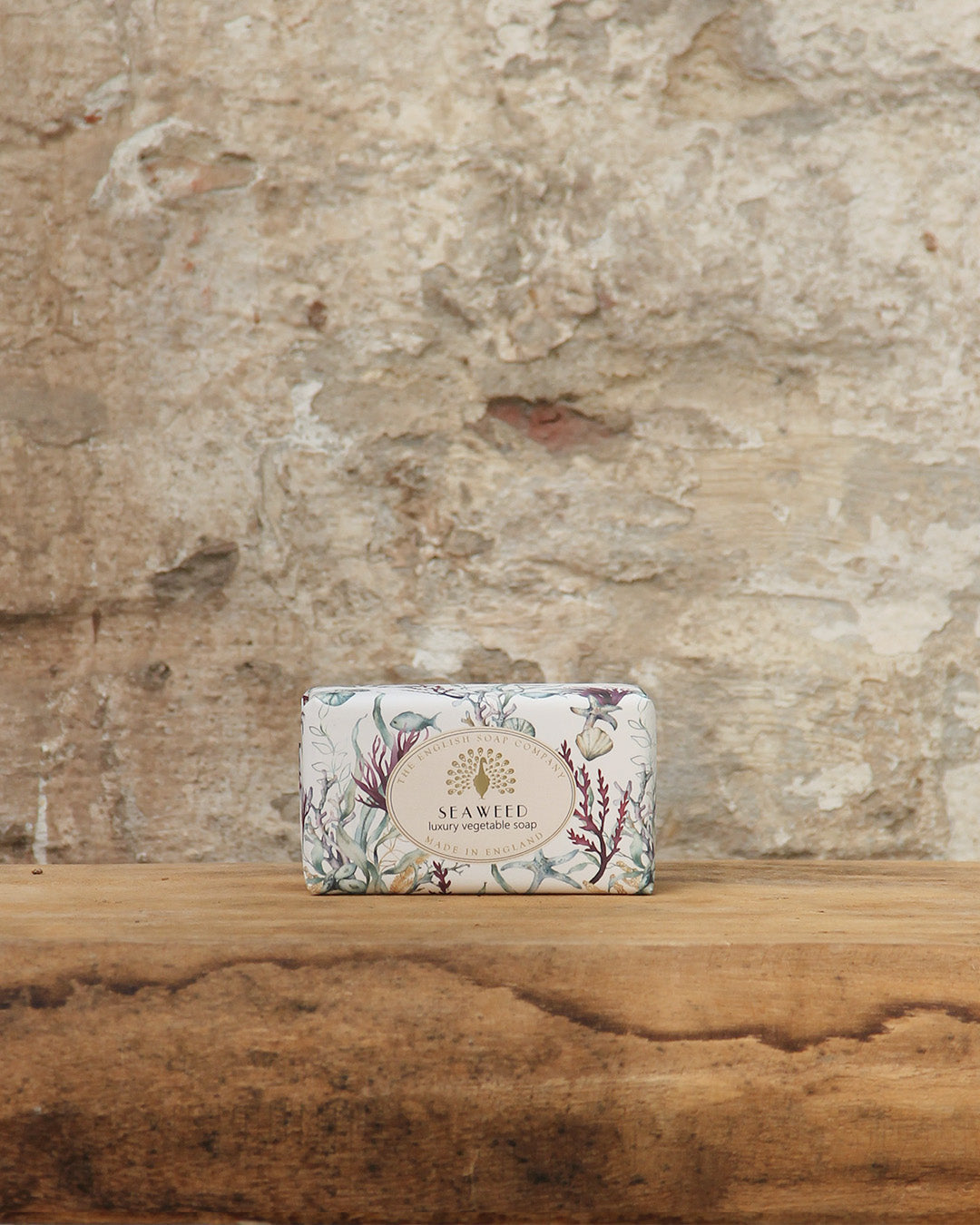 Ein Stück The English Soap Company ESC | 'Seaweed' pflanzliche Handseife aus Bio-Seetang in einer blumigen Verpackung ruht auf einer hölzernen Oberfläche mit einer strukturierten, verwitterten Steinwand im Hintergrund.