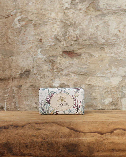 Ein Stück The English Soap Company ESC | 'Seaweed' pflanzliche Handseife aus Bio-Seetang in einer blumigen Verpackung ruht auf einer hölzernen Oberfläche mit einer strukturierten, verwitterten Steinwand im Hintergrund.