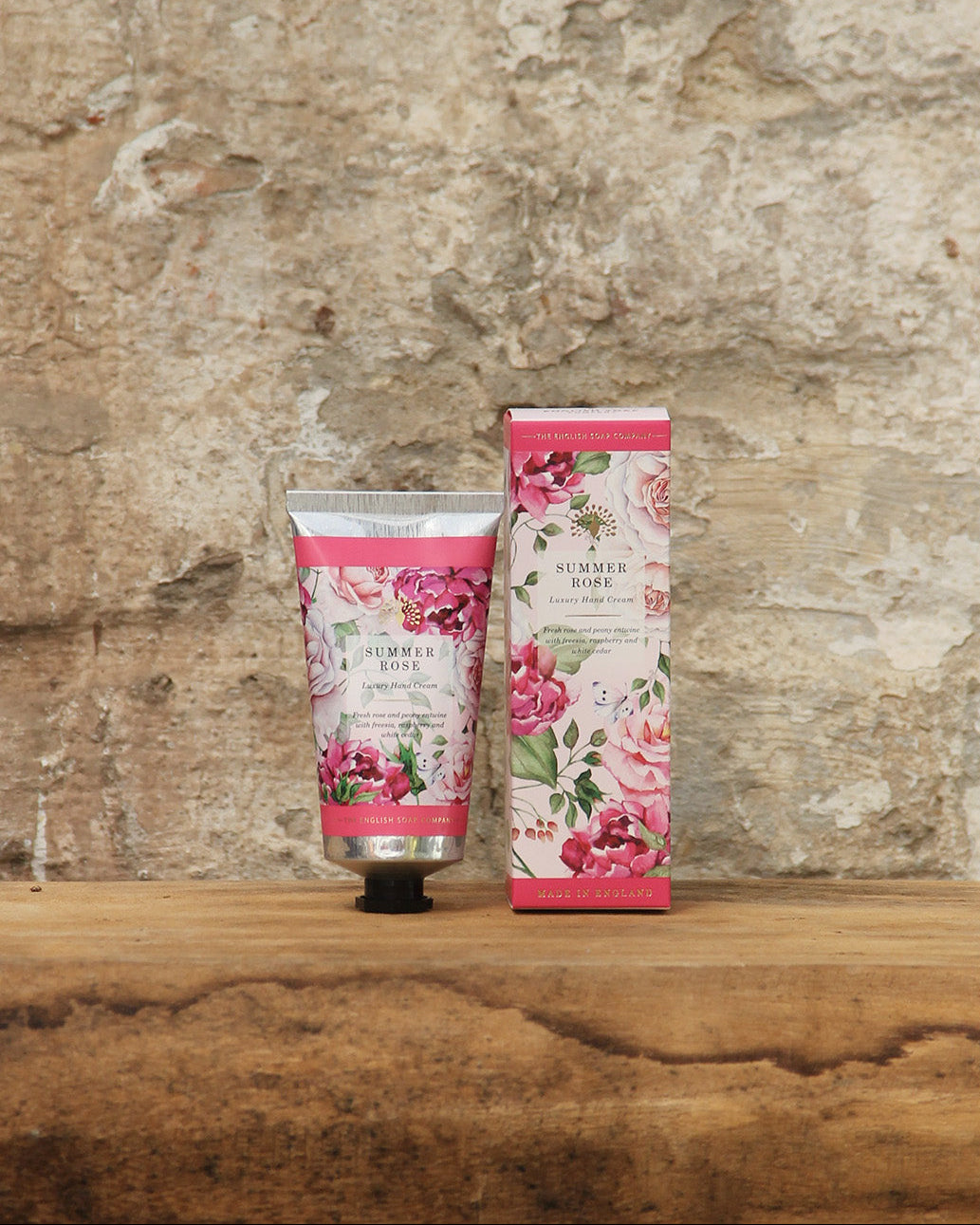 Eine blumige ESC | 'Summer Rose' Handcreme 75 ml von The English Soap Company und die dazugehörige Box ruhen auf rustikalem Holz auf Stein, mit natürlichen Inhaltsstoffen und plastikfreier Verpackung.