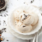 Drei Kugeln cremiges Eis in einer weißen Schale, übergossen mit L.W.C. Michelsen Gourmet-Sirup „Earl Grey“ und garniert mit losen schwarzen Teeblättern, wobei weitere Teeblätter auf einer weißen Holzfläche daneben verstreut liegen.