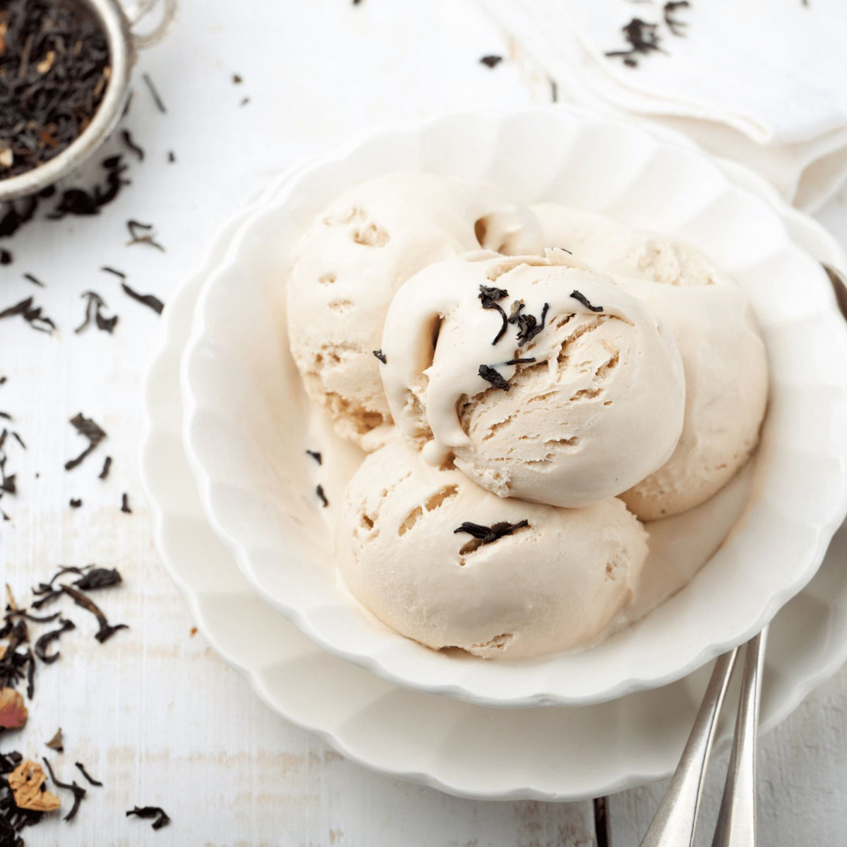 Drei Kugeln cremiges Eis in einer weißen Schale, übergossen mit L.W.C. Michelsen Gourmet-Sirup „Earl Grey“ und garniert mit losen schwarzen Teeblättern, wobei weitere Teeblätter auf einer weißen Holzfläche daneben verstreut liegen.
