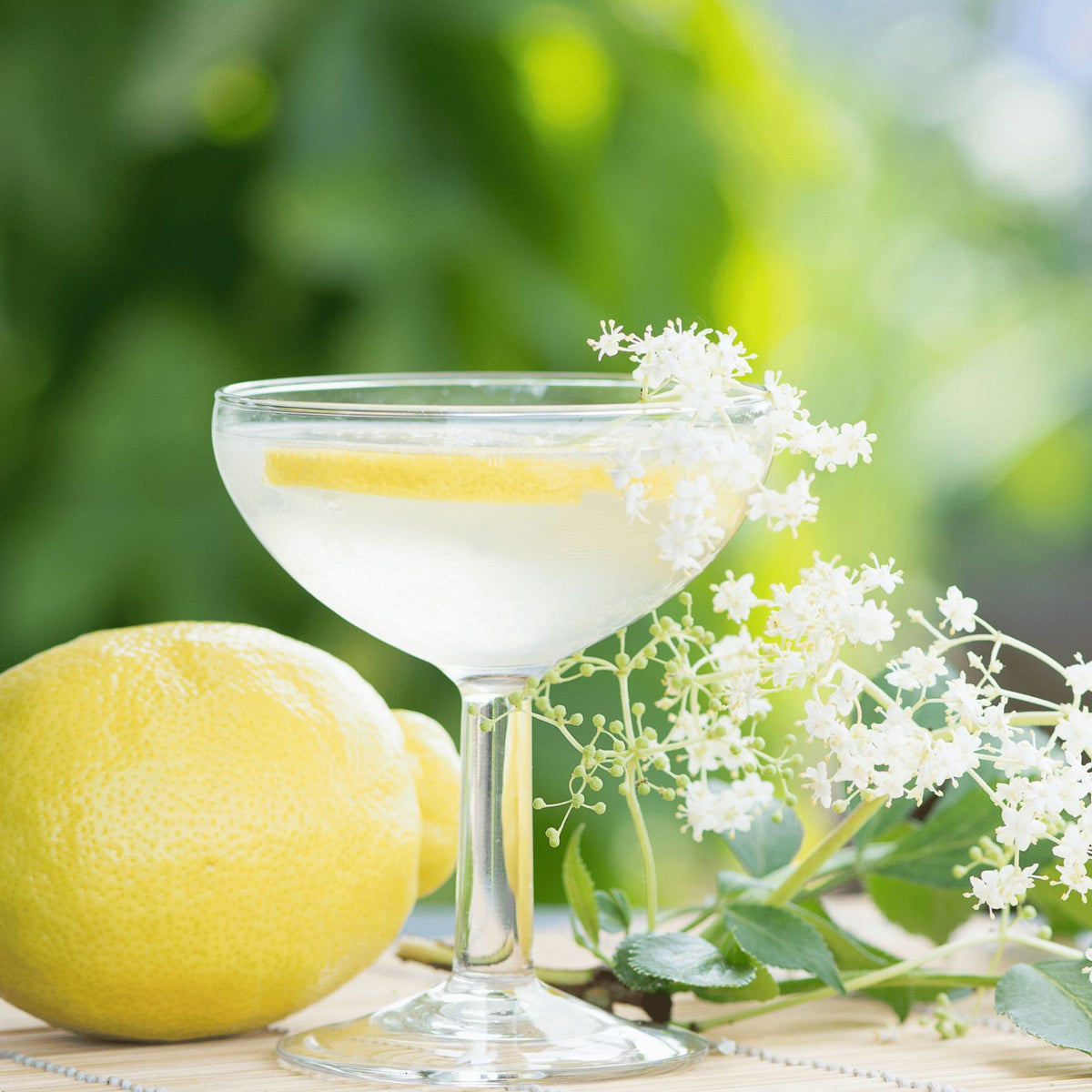 Ein mit einer Zitronenscheibe garniertes Cocktailglas, das neben einer ganzen Zitrone und zarten weißen Holunderblüten auf einer Holzunterlage vor einem sanft unscharfen grünen Hintergrund steht, unterstreicht auf wunderschöne Weise den erfrischenden Charakter von L.W.C. Michelsen | Gourmet Sirup Holunderblüte 250 ml.