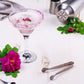 Ein rosa Cocktail in einem Margarita-Glas mit gezuckertem Rand, garniert mit Blütenblättern, steht auf einem Tisch neben einer rosa Rose, einem Cocktailshaker und einer Eiszange. Die zarten blumigen Noten des L.W.C. Michelsen Gourmet Sirup Rose verleihen dem Getränk eine elegante Süße, die es ebenso bezaubernd im Geschmack wie für das Auge macht.
