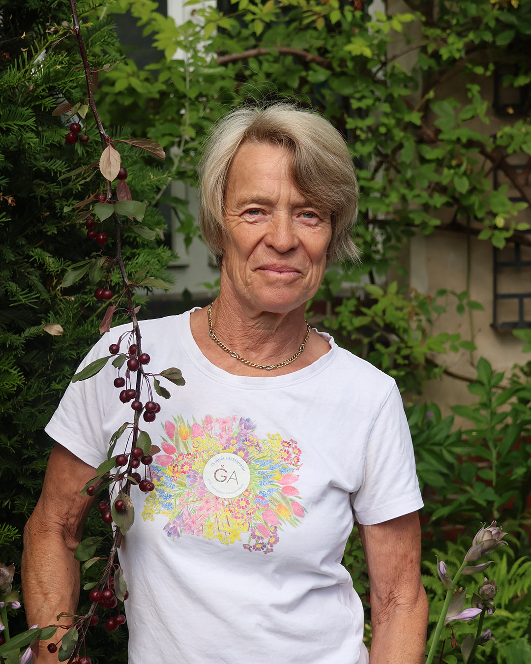 Eine ältere Frau mit kurzen blonden Haaren steht in einem üppigen Garten, nachdem sie den Workshop "Rosenschnitt einfach erklärt" der Königlichen Gartenakademie besucht hat. Sie trägt ein weißes T-Shirt mit einem Blumenmuster und eine goldene Halskette, im Hintergrund sind Grünpflanzen und Beeren zu sehen.