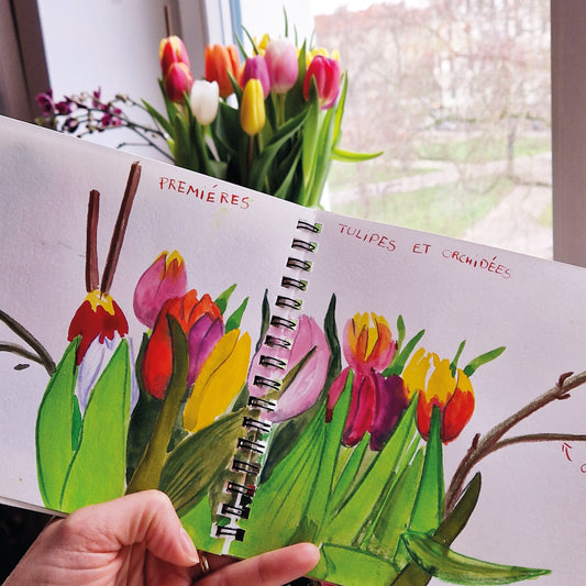 Eine Hand hält ein Skizzenbuch mit lebendiger Tulpen- und Orchideenkunst, gemalt mit Aquarellfarben, vor blühenden Tulpen. Auf dem Skizzenbuch steht "Premières Tulipes et Orchidées". Produkt: Fr. 24.04.2026 | 10-13 Uhr MIXED-MEDIA-WORKSHOP Mein Garten in einem Tagebuch, Königliche Gartenakademie.