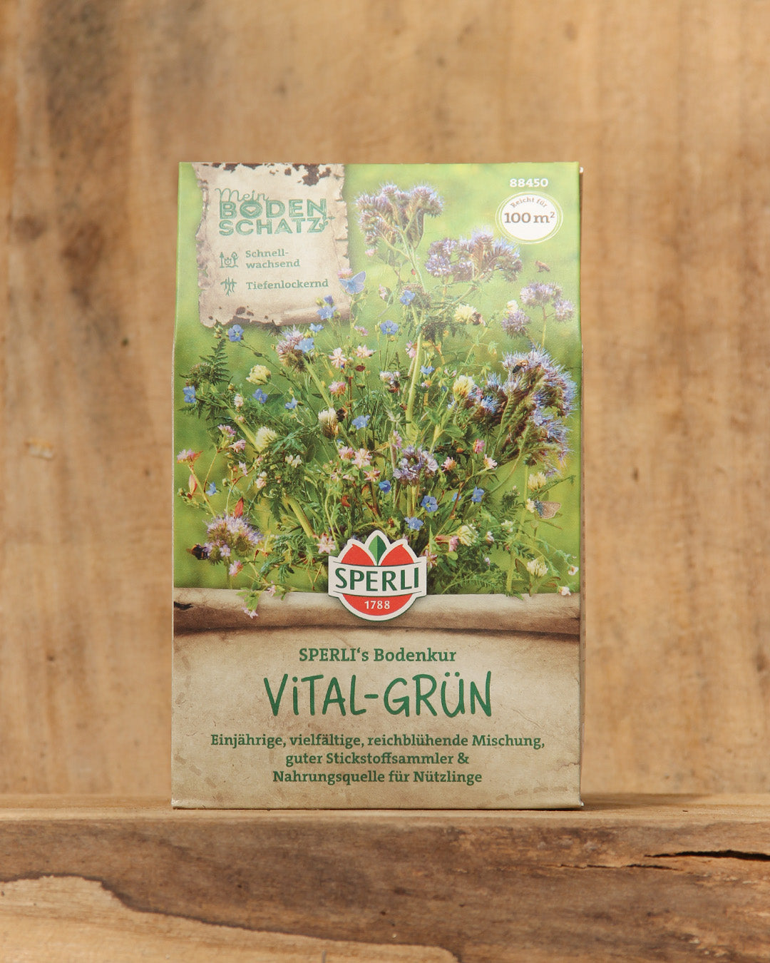 Eine 450-g-Packung der Sperli's Gründüngermischung Bodenkur 'Vital-Grün' steht aufrecht auf einer Holzfläche und zeigt den Markennamen und die Produktangaben in deutscher Sprache mit abgebildeten blühenden Pflanzen vor einem verschwommenen Holzhintergrund.