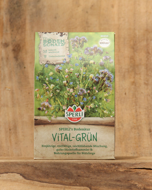 Eine 450-g-Packung der Sperli's Gründüngermischung Bodenkur 'Vital-Grün' steht aufrecht auf einer Holzfläche und zeigt den Markennamen und die Produktangaben in deutscher Sprache mit abgebildeten blühenden Pflanzen vor einem verschwommenen Holzhintergrund.