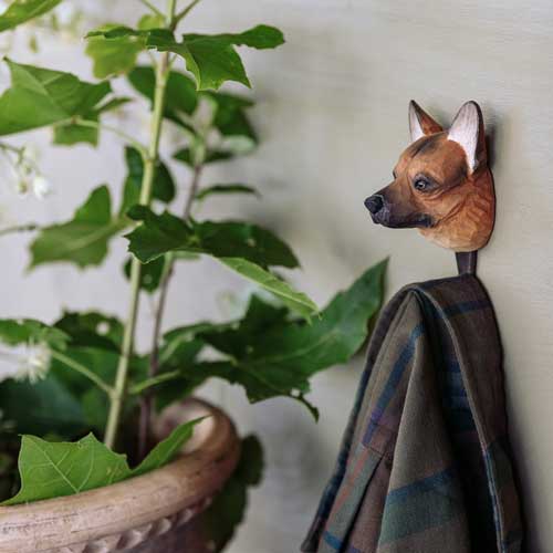 Der handgeschnitzte Wandhaken aus Lindenholz 'Deutscher Schäferhund' von Wildlife Garden hält ein kariertes Halstuch und hängt neben einer Pflanze mit grünen Blättern und kleinen weißen Blüten.