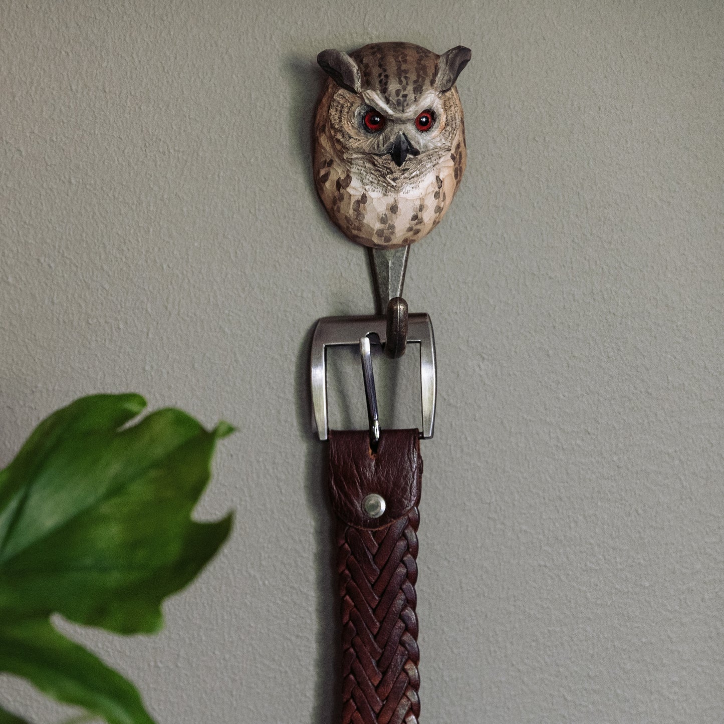 Ein geflochtener brauner Ledergürtel hängt am Wildlife Garden | 'Uhu' handgeschnitzter Wandhaken aus Lindenholz, geformt wie ein Eulenkopf. In der unteren linken Ecke ist ein grünes Pflanzenblatt vor einer hellgrauen Wand zu sehen.
