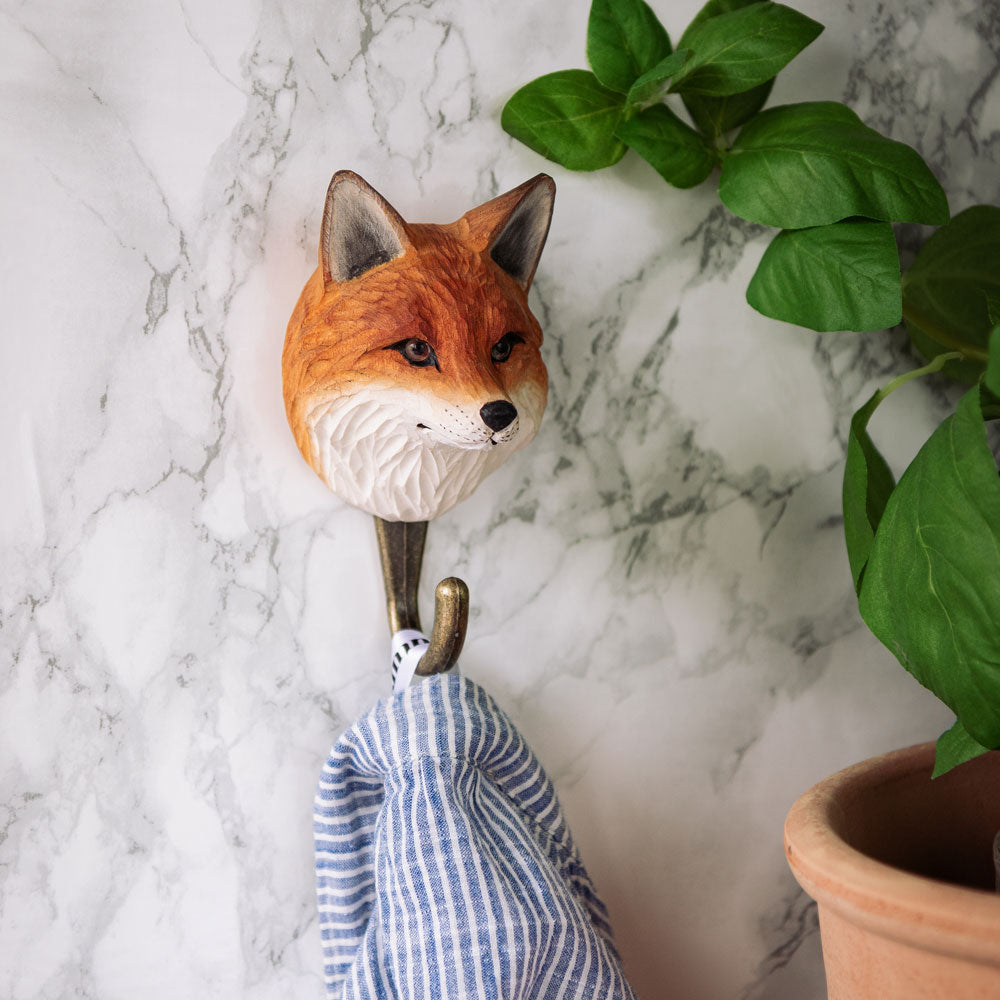 Der Wildlife Garden Handgeschnitzter Wandhaken aus Holz 'Fuchs' hält ein blau-weiß gestreiftes Tuch mit grünen Blättern und einem Terrakottatopf in der Nähe, alles vor einem weißen Hintergrund mit Marmormuster.