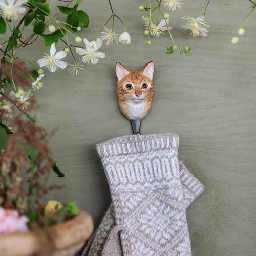 Der Wildlife Garden Handgeschnitzter Wandhaken aus Holz 'Hauskatze' von Wildlife Garden ist ein gemusterter Strickfäustling mit weißen Blüten und grünen Blättern darüber - ein charmantes Katzen Deko und Geschenk für Naturliebhaber vor grüner Wand.
