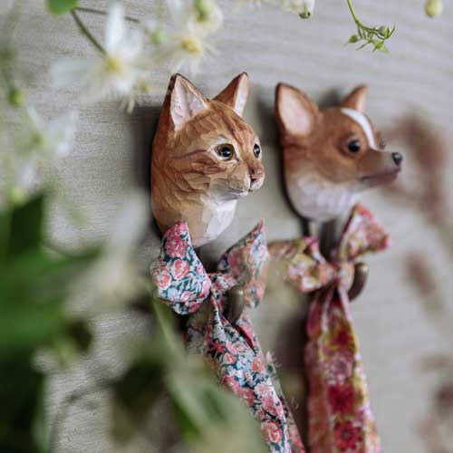 Zwei handgeschnitzte Wildlife Garden Wandhaken aus Holz 'Hauskatze', in Form einer orangefarbenen Katze und eines braunen Hundes mit floralen Schleifen-einzigartige Katzen Deko von Wildlife Garden, ideal als Geschenk Naturliebhaber, mit verschwommenen Blumen und Pflanzen im Vordergrund.