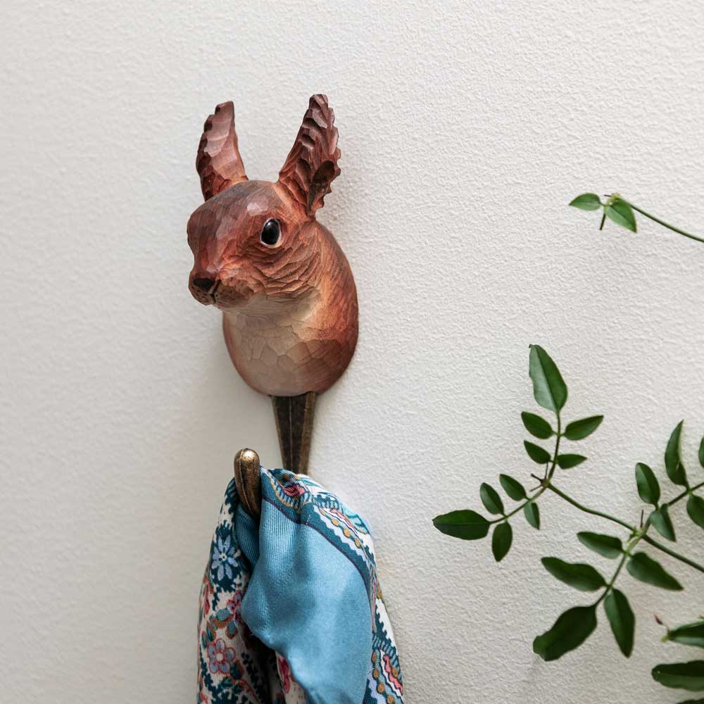 Der handgeschnitzte Wandhaken aus Holz 'Eichhörnchen' von Wildlife Garden hält ein blau gemustertes Tuch. Daneben steht eine grüne Blattpflanze vor einer weißen, strukturierten Wand.