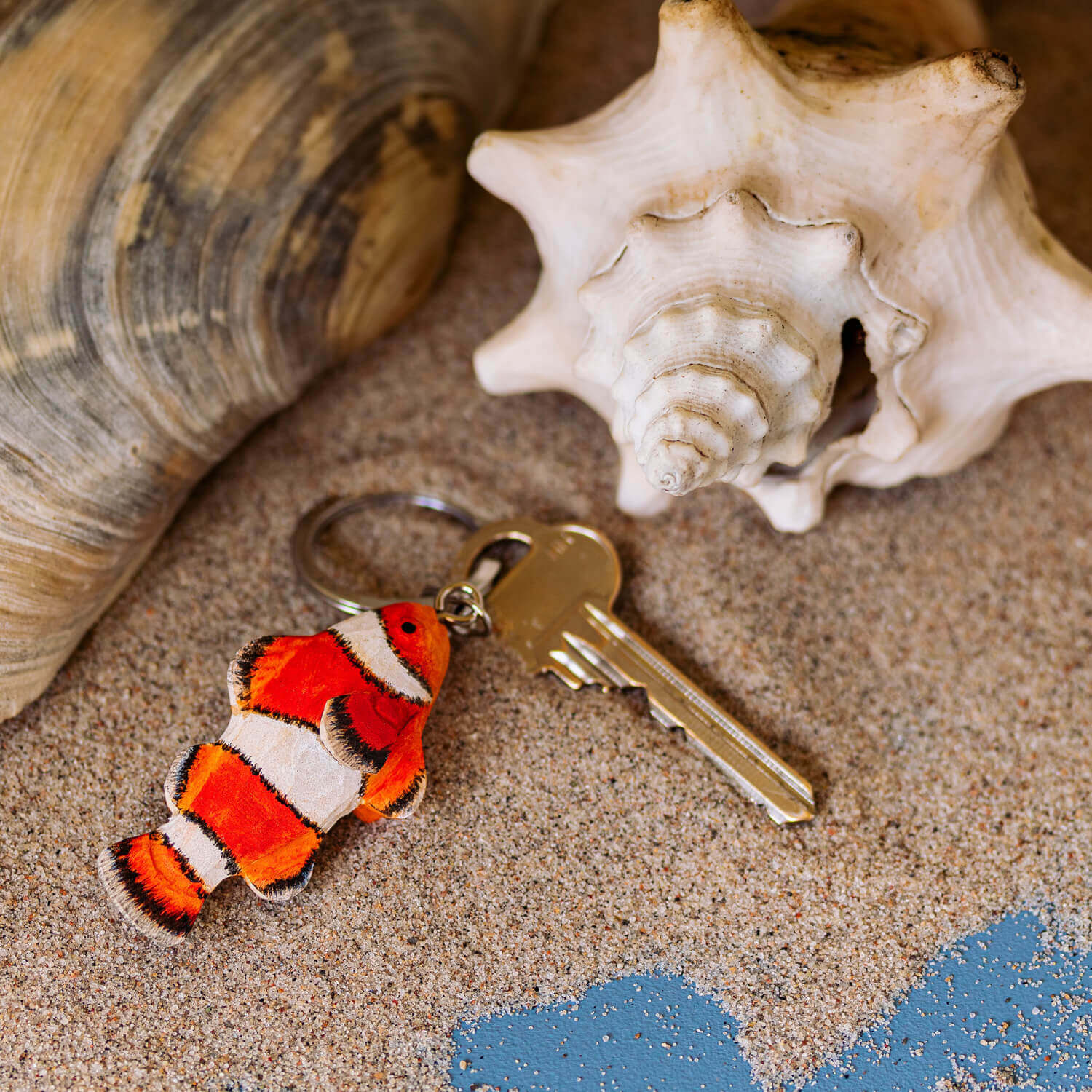 Ein handgefertigter Schlüsselanhänger aus Holz | Clownfisch von Wildlife Garden liegt neben Muscheln im Sand - ein charmantes Geschenk für Meereswelt-Fans.