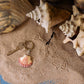 A Wildlife Garden Handgefertigter Schlüsselanhänger aus Holz Muschel liegt auf sandigem Boden neben großen Muscheln und Vogelspuren - ein charmantes maritimes Accessoire von Wildlife Garden.