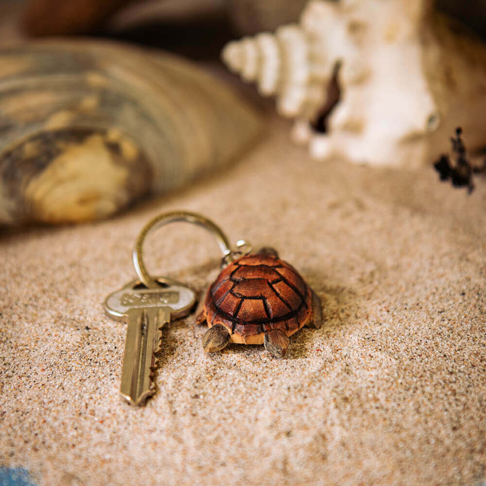 Ein Metallschlüssel mit dem Wildlife Garden Schildkröte Schlüsselanhänger aus Holz ruht auf Sand, große Muscheln dahinter. Dieser handgefertigte Schildkröten Schlüsselanhänger von Wildlife Garden fügt Ihrer Meeresschildkröte Accessoire Sammlung Charme hinzu.