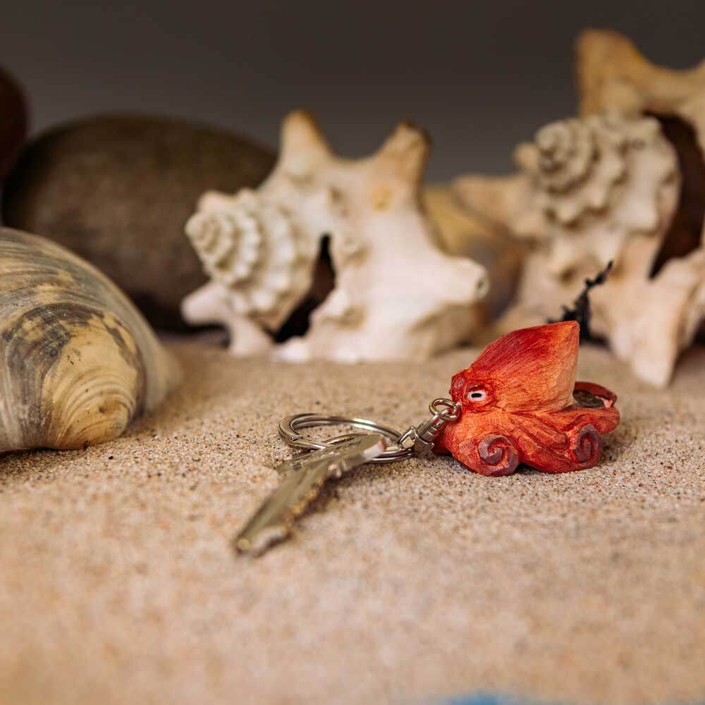 A Wildlife Garden Handgefertigter Schlüsselanhänger aus Holz in Oktopus-Form und ein Metallschlüssel liegen auf sandigem Boden, umgeben von Muscheln und Steinen - ein charmantes maritimes Geschenk.