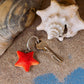 Ein Wildlife Garden | Handgefertigter Schlüsselanhänger aus Holz | Seestern von Wildlife Garden ruht auf sandigem Boden neben einem Metallschlüssel und zwei Muscheln, eine spiralförmig und eine muschelartig.
