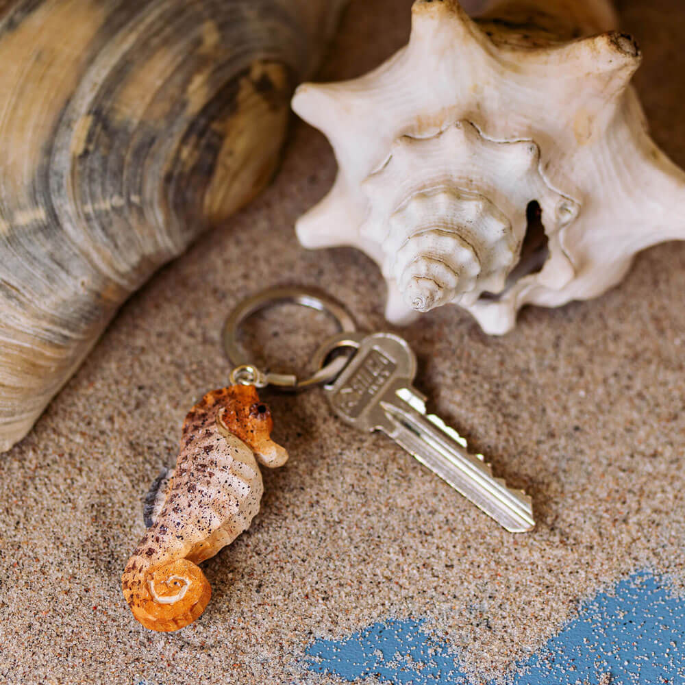 Der Wildlife Garden Handgefertigter Schlüsselanhänger aus Holz Seepferd liegt mit einem silbernen Schlüssel auf Sand neben einer großen Muschel - das perfekte maritime Accessoire für Strandliebhaber von Wildlife Garden.