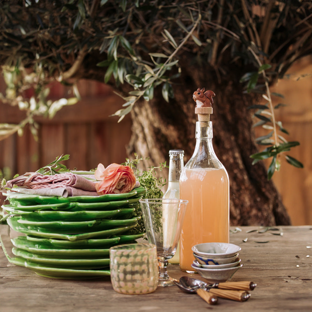 Auf einem rustikalen Tisch im Freien unter einem Olivenbaum stapeln sich grüne Teller, gefaltete Servietten mit einer rosafarbenen Blume, Gläser, Schüsseln, Besteck und Flaschen mit rosafarbener Limonade, die mit dem handgeschnitzten Flaschenverschluss Holz 'Tannenzapfen' von Wildlife Garden verziert sind.