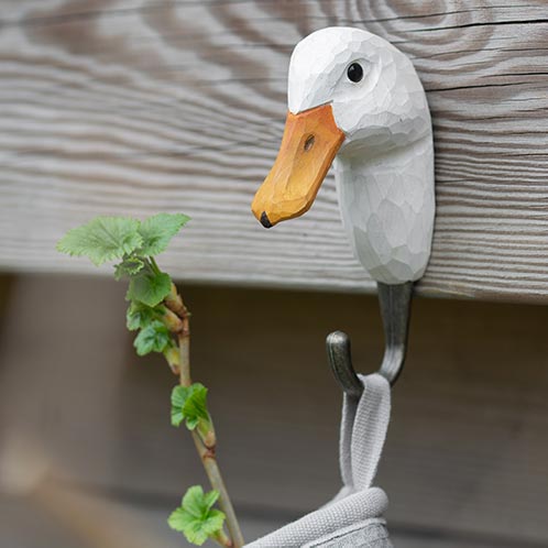 Der handgeschnitzte Wandhaken aus Holz 'Ente' von Wildlife Garden ist an einer Holzwand befestigt und hält einen grauen Gurt sowie einen grünen Blattstiel.
