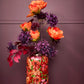 Ein farbenfrohes Blumenarrangement mit orangefarbenen und violetten Blüten ist in der Vase „Bloom“ aus Metall in Rosarot (30 x 15 cm) von 2 Have zu sehen, die sich durch ein dekoratives Blumenmuster auszeichnet. Die Vase steht auf einer Holzunterlage vor einer violetten Wand.