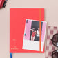 Ein Tinne Mia | Notizbuch mit japanischem Verschluss 'Cinnamon' hält ein Foto von zwei Personen an einer rosa Wand. In der Nähe befinden sich eine Sonnenbrille auf einem gestreiften Etui und ein Teil einer rosa Metalltasche, alles auf einer hellbeigen Fläche.