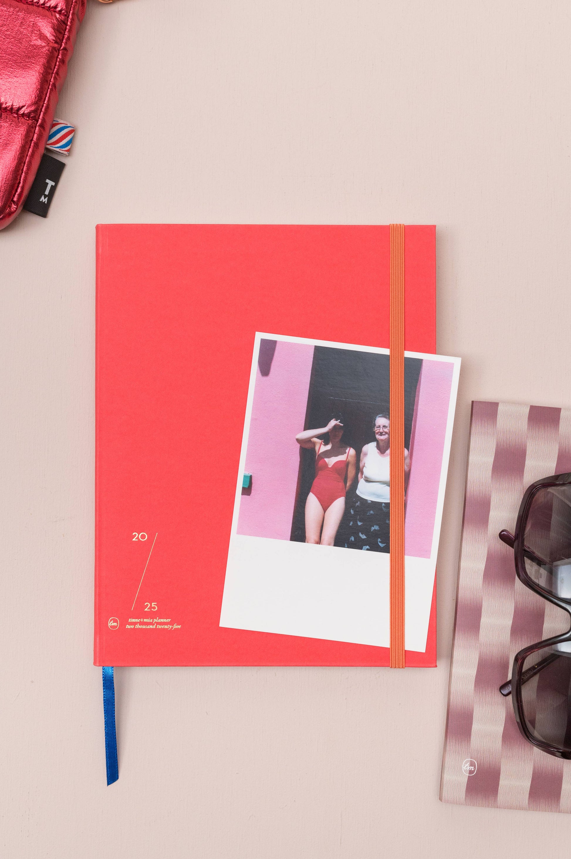 Ein Tinne Mia | Notizbuch mit japanischem Verschluss 'Cinnamon' hält ein Foto von zwei Personen an einer rosa Wand. In der Nähe befinden sich eine Sonnenbrille auf einem gestreiften Etui und ein Teil einer rosa Metalltasche, alles auf einer hellbeigen Fläche.