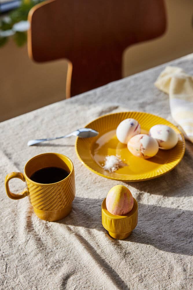 Ein Kaffeebecher Gelb, 2-teilig, der Sagaform GmbH, gefüllt mit Kaffee, steht auf einem beigen Tischtuch im Sonnenlicht, daneben ein kleiner Eierbecher und ein gelber Teller mit gefärbten Eiern - skandinavisches Design vom Feinsten. Dahinter stehen ein Stuhl und eine Serviette.