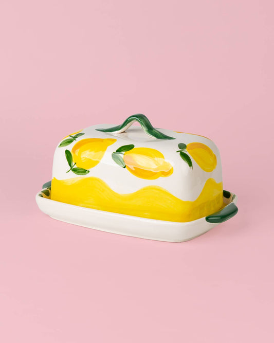 Die handgefertigte Keramik-Butterdose „Limone“ in Gelb und Weiß von Alfar Tierra Cocida ist mit gelben Zitronen und grünen Blättern verziert und steht auf einem hellrosa Hintergrund. Das mediterrane Geschirr besticht durch grüne Griffe und ein wellenförmiges, gelbes Muster.