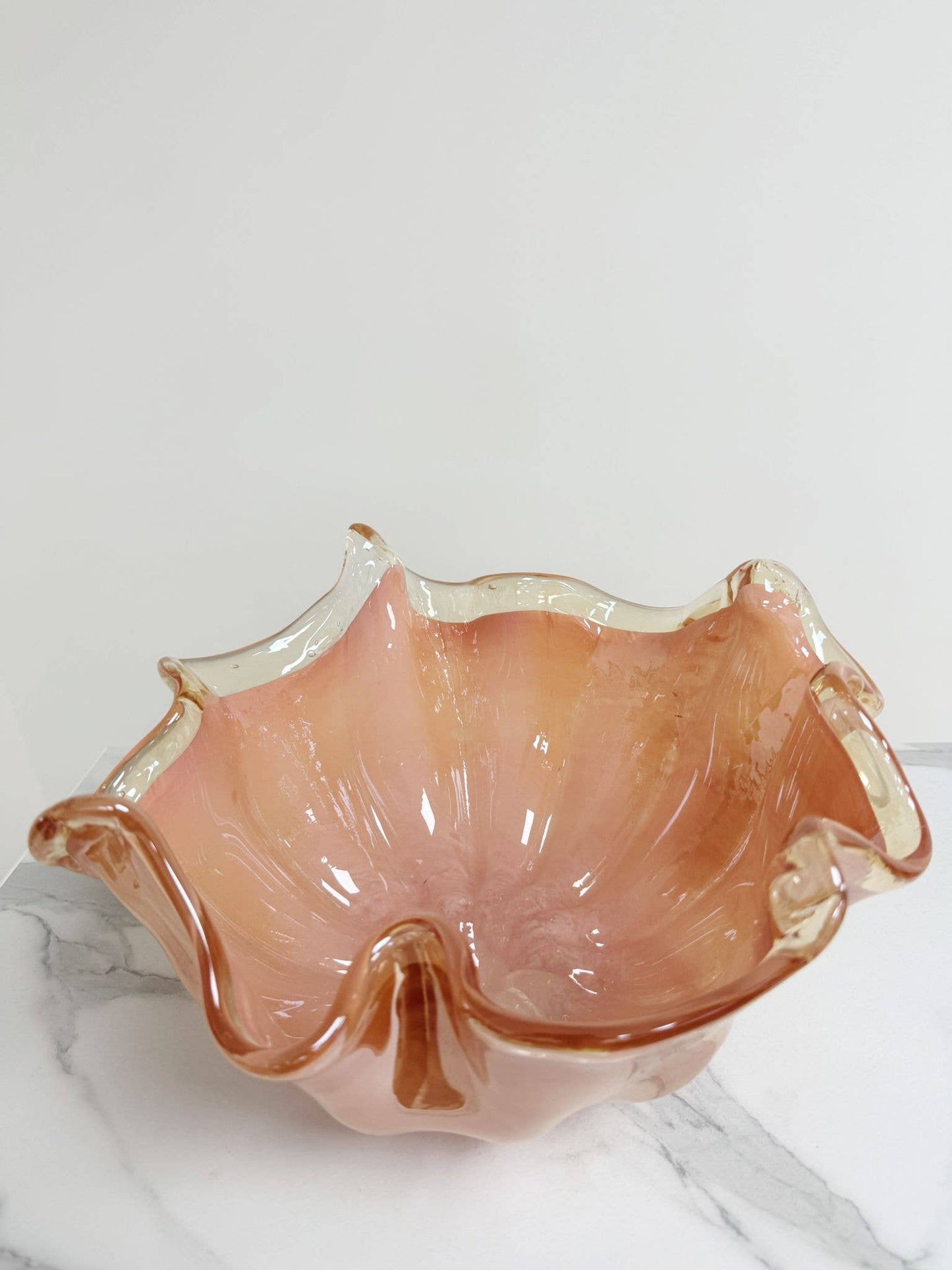 Die rosa Dekoschale „Fazzoletto“ aus Murano-Glas (H 20, Ø 22 cm) von Murano Dream glänzt mit ihrem gewellten Rand auf weißem Marmor und verkörpert die Eleganz der italienischen Glaskunst.