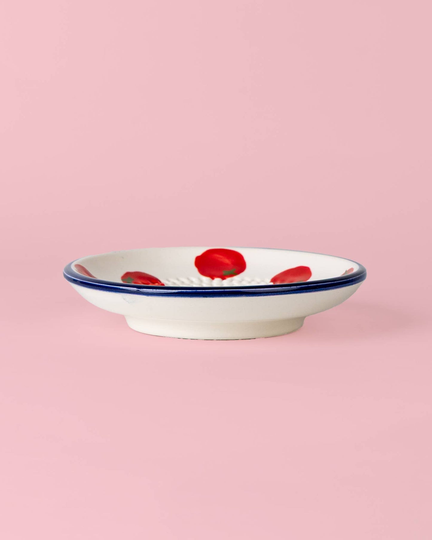 Eine flache weiße Keramikschale mit blauem Rand und rot gemalten Blumen: die „Pomodoro“ von Alfar Tierra Cocida | Kleine Keramik Knoblauchreibe, handgefertigt von Alfar Tierra Cocida in Spanien, vor einem hellrosa Hintergrund.