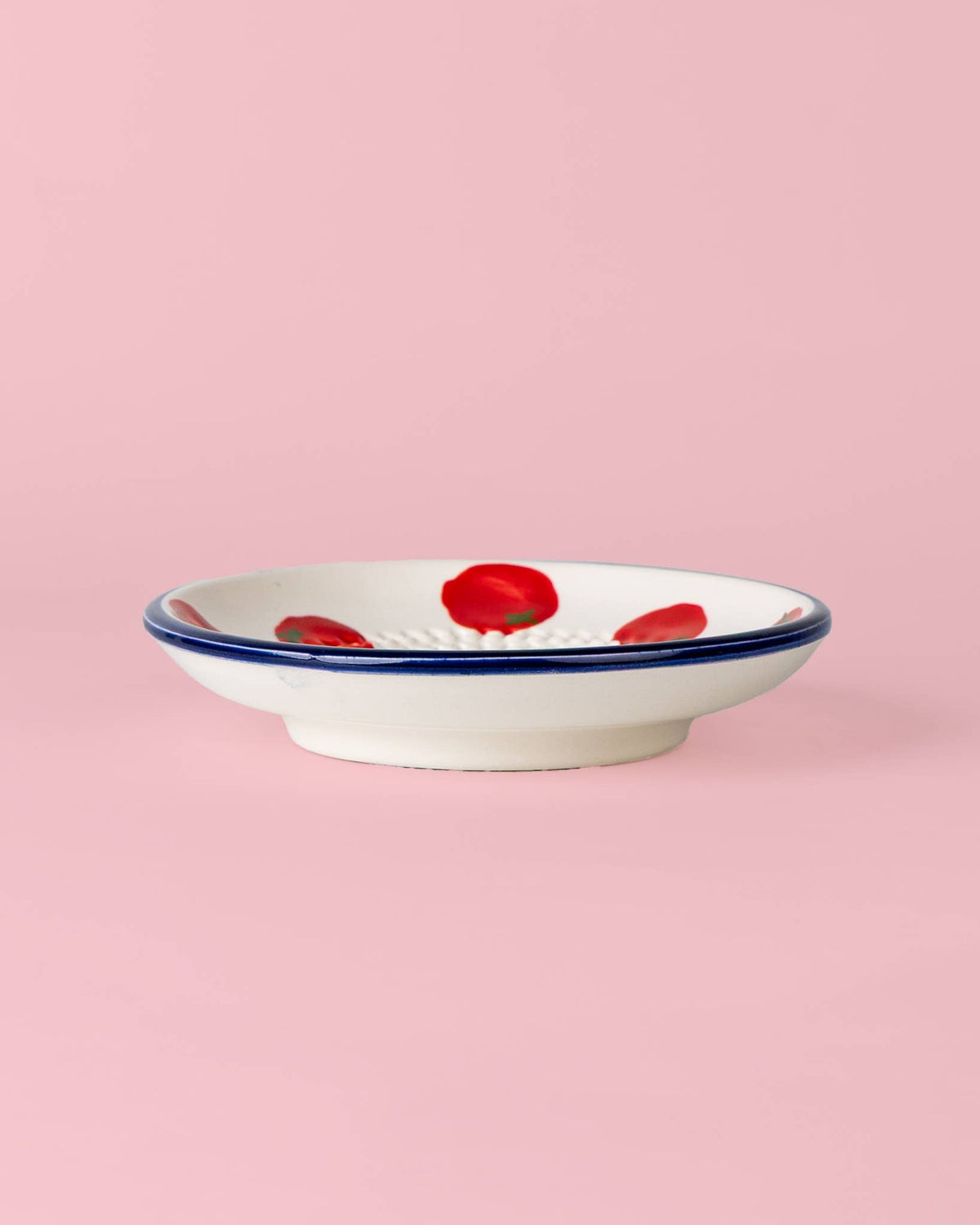 Eine flache weiße Keramikschale mit blauem Rand und rot gemalten Blumen: die „Pomodoro“ von Alfar Tierra Cocida | Kleine Keramik Knoblauchreibe, handgefertigt von Alfar Tierra Cocida in Spanien, vor einem hellrosa Hintergrund.