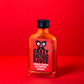 Eine quadratische Glasflasche mit Crazy Bastard Sauce Trinidad Scorpion & Clementine 100 ml scharfe Soße steht auf einer roten Fläche mit rotem Hintergrund. Das Etikett zeigt stilisierte Augen, Text und Schärfesymbole für Chilischoten am unteren Rand.