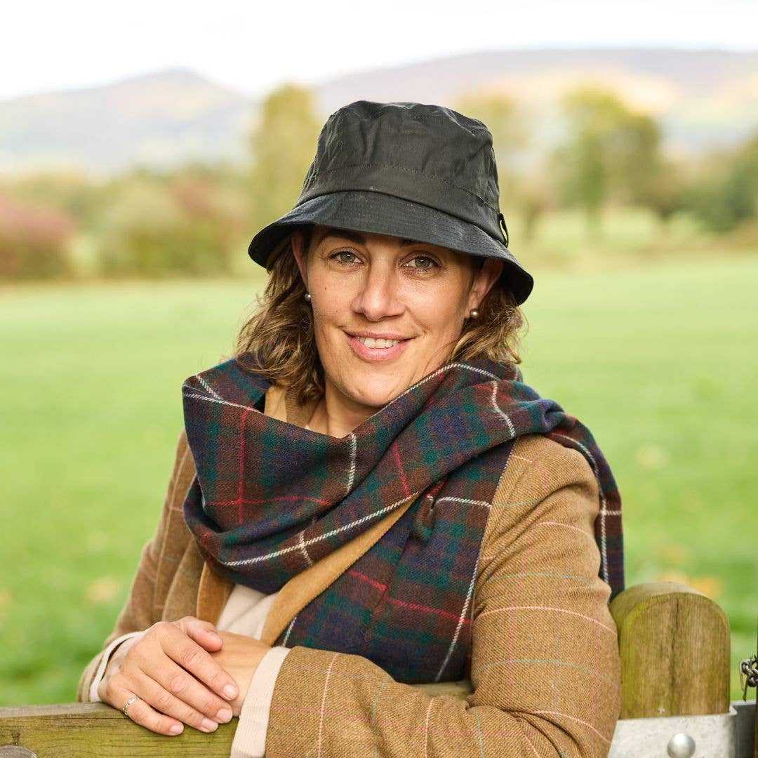 Eine Frau, die einen schwarzen Krempenhut „Wachstropfen“ von Tweedmill Textiles, einen braunen karierten Schal und einen hellbraunen Mantel trägt, lächelt, während sie sich auf einem Holzzaun in einem grünen Feld anlehnt; im Hintergrund sind verschwommene Bäume und Hügel zu sehen.