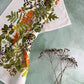 Ein Geschirrtuch aus 100 % Bio-Baumwolle mit einem herbstlichen Botanical-Print von Koustrupco liegt auf einer grünen Fläche neben einem Zweig mit getrockneten braunen Blumen.