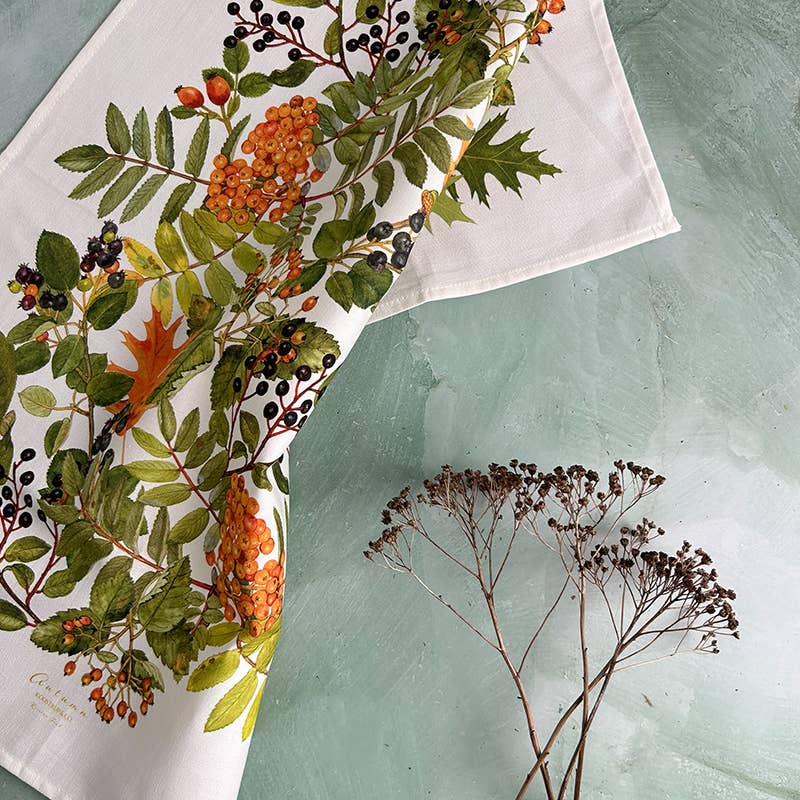 Ein Geschirrtuch aus 100 % Bio-Baumwolle mit einem herbstlichen Botanical-Print von Koustrupco liegt auf einer grünen Fläche neben einem Zweig mit getrockneten braunen Blumen.