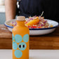 Eine 200-ml-Flasche Yiayia and friends Weißer Balsamico-Essig, verziert mit blauen Kreisen und verspielten Augen, steht vor einem bunten Salat, der ein reines Dressing suggeriert. Im Hintergrund ist eine Person in einer dunklen Schürze zu sehen.