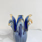Der Murano-Traum | Die mittelgroße „Giusy“-Vase aus blauem Murano-Glas (H 24, Ø 19 cm) mit wellenförmigem, blütenähnlichem Rand in Blau und Bernstein präsentiert edle italienische Glaskunst auf weißem Marmor vor hellem Hintergrund.
